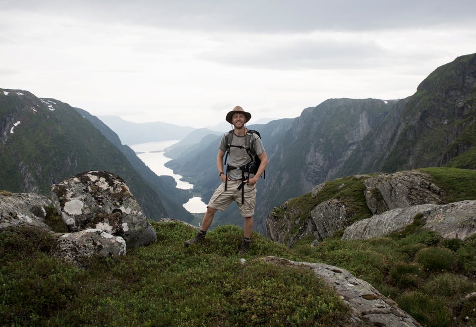 Stilig mann i knalltøff hatt på fjell med fjord bak seg.