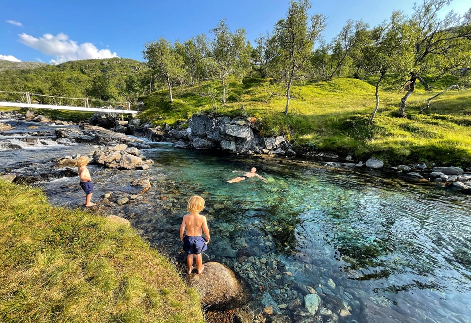 Bading i badekulp, en voksen mann bader og to unge gutter står ved elvekanten og ser på.