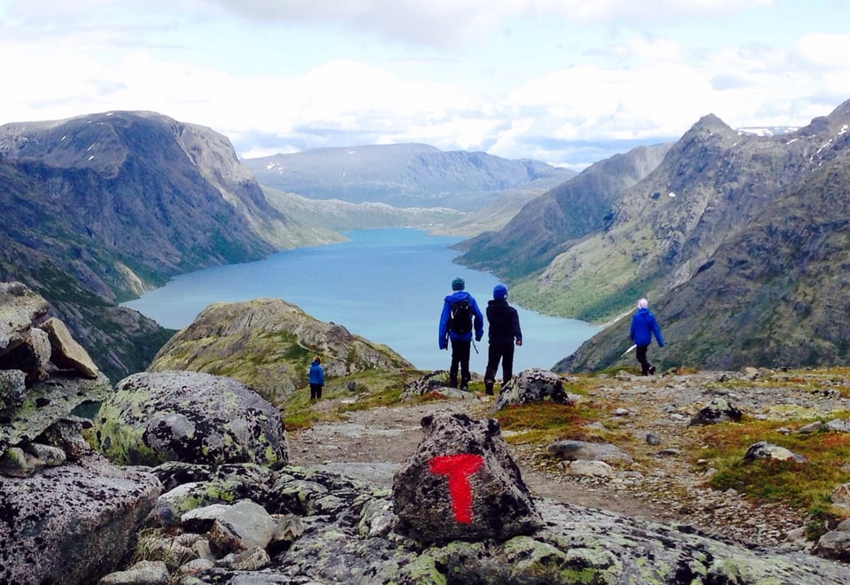 Personer og T-merket stein på fjelltopp, med utsikt mot vann og fjell i bakgrunnen
