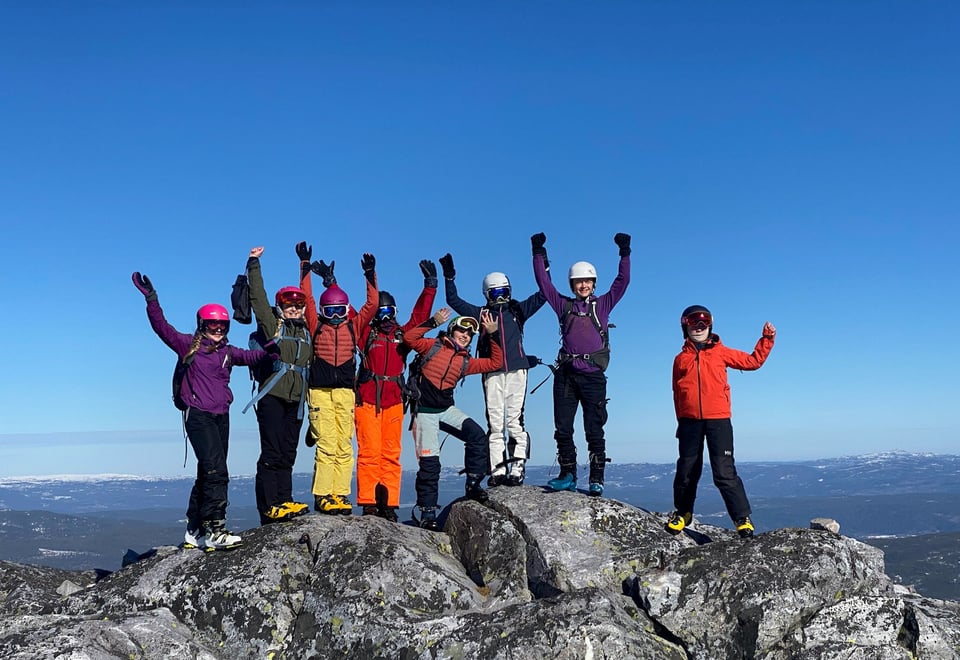 Ungdommer i skiutstyr står på toppen av et fjell med hendene i været