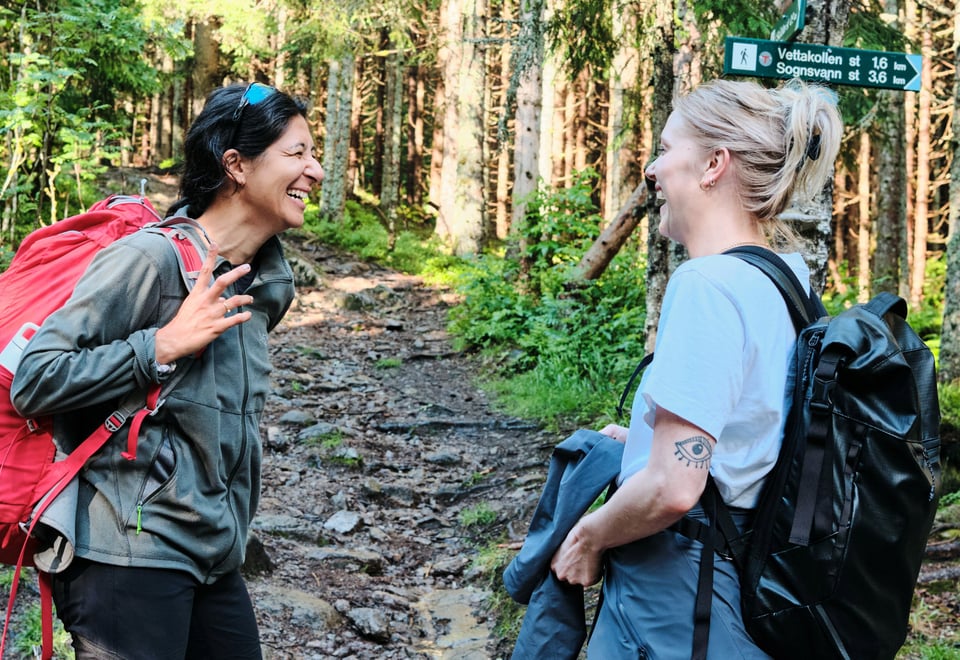 To personer smilende og pratsomme på en skogsti i Oslomarka. Begge bærer tursekker, og i bakgrunnen sees et skilt som viser retningene til Vettakollen (1,6 km) og Sognsvann (3,6 km)