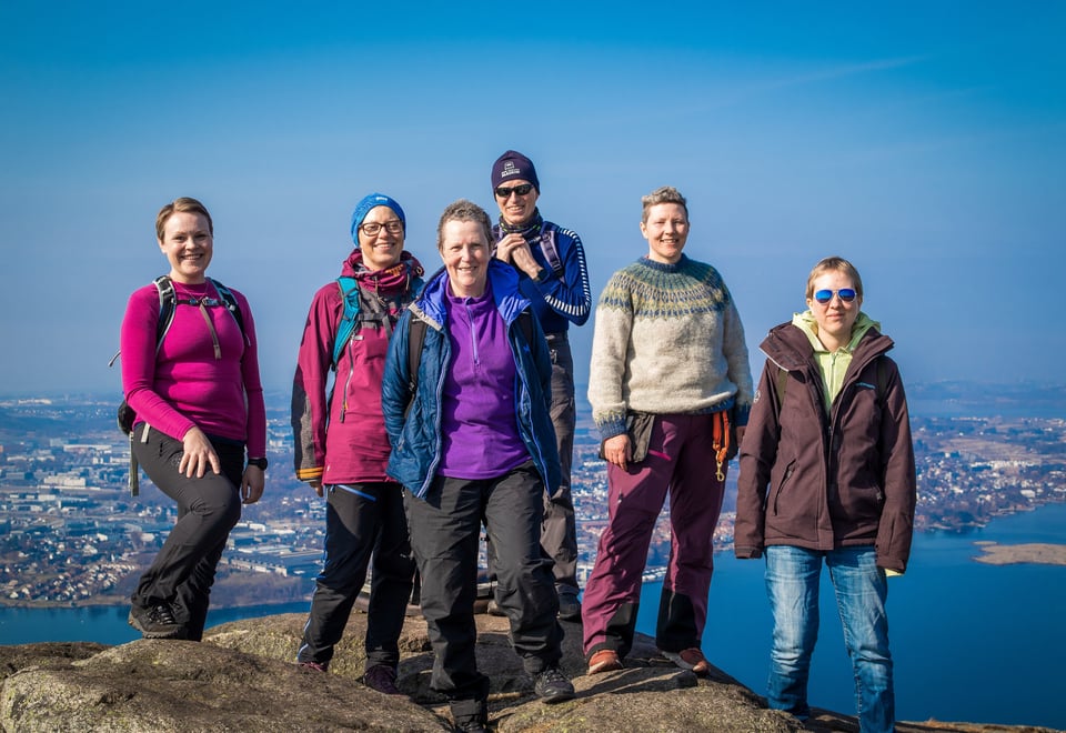 Seks personer står på toppen av et fjell og ser i kamera. Utsikten bak dem viser sjø og bebyggelse.