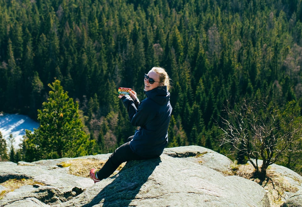 En kvinne sitter på en stein ute i naturen med en skogkledd åsside i bakgrunnen. Hun smiler mens hun holder en Kvikk Lunsj-sjokolade i hånden og har på seg solbriller og en mørk turjakke. Det ser ut som hun nyter en pause på en tur i skogen