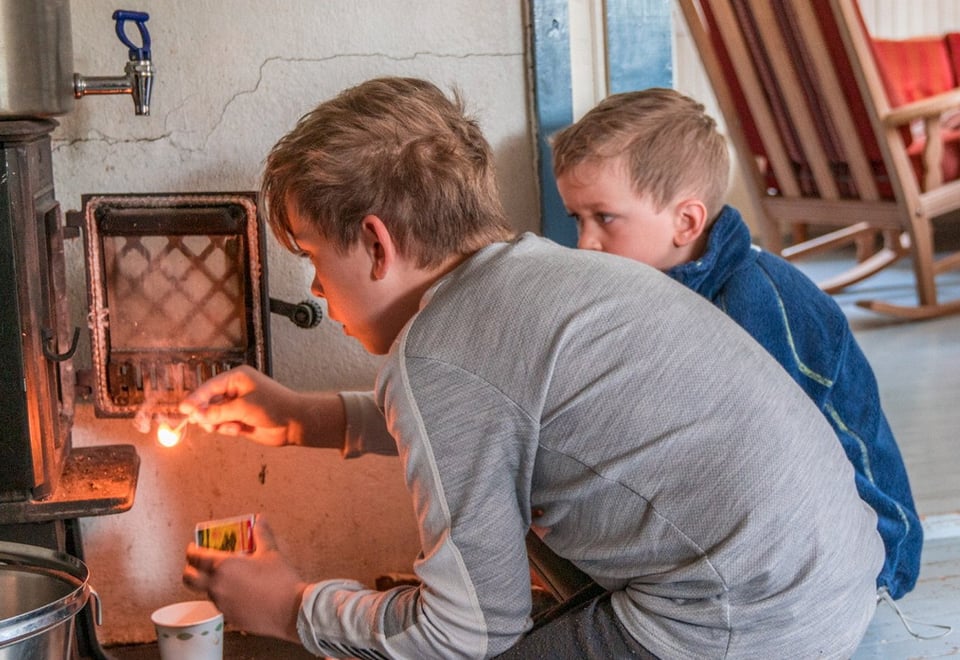 To gutter som fyrer i en vedovn. Den ene gutten har en fyrstikk i hånden.