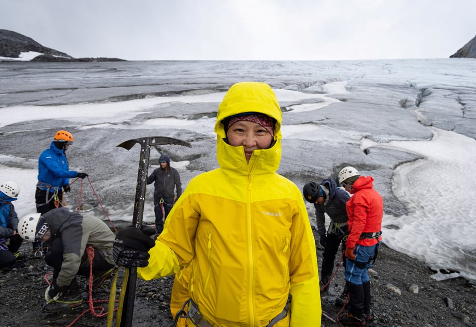 Mongolsk kvinne i gul jakke og isøks i hånden smiler i kamera foran isbre.