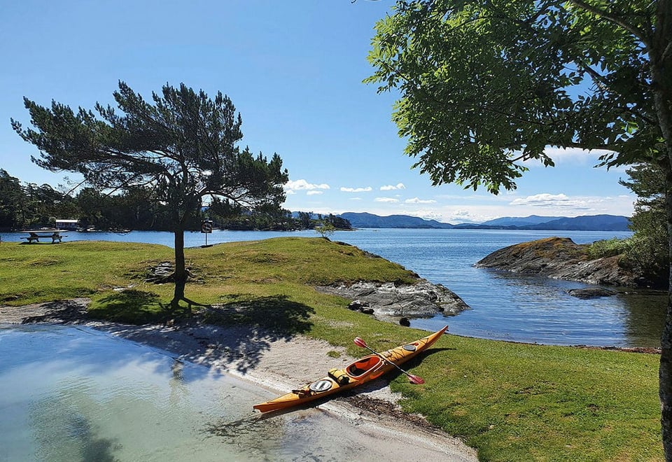 Kajakk ligger på sandstrand i idyllisk frilkuftsområde med sol, sjø og grønn gressplen.