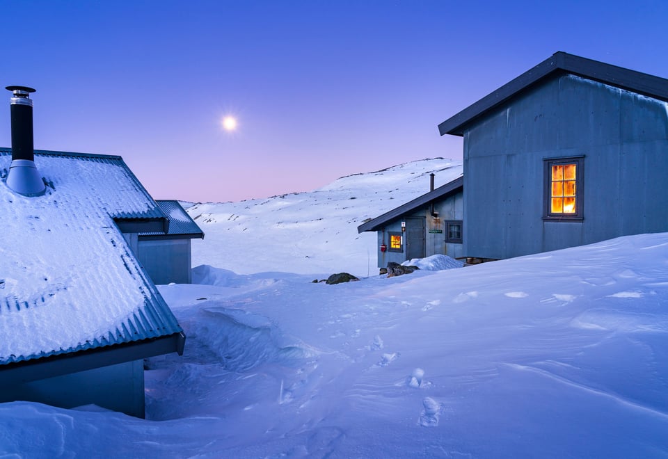 Et hyttetun omgitt av snø og måneskinn. Hyttene er i grå farge og himmelen er nærmest lilla.