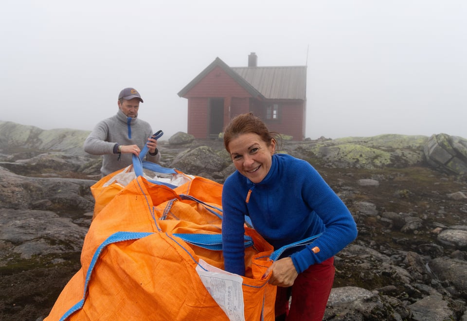 Kvinne i blå genser smiler foran hytte i tåkeheim.