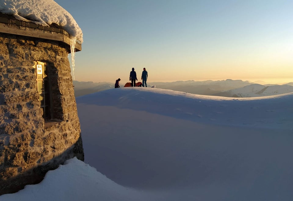 Tre personer står på snøkledd fjelltopp i solnedgang med hytten til venstre i bildet nærmere kamera.