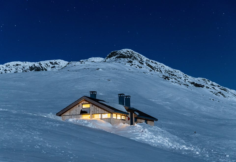 Hytte i vakkert snølandskap i fjellet med lys inne og stjernehimmel over hytta.