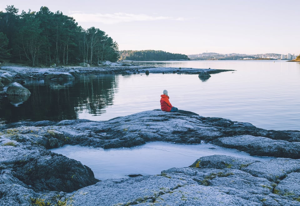 bilde av en person i oransj jakke som sitter på svaberg og ser utover vannet.