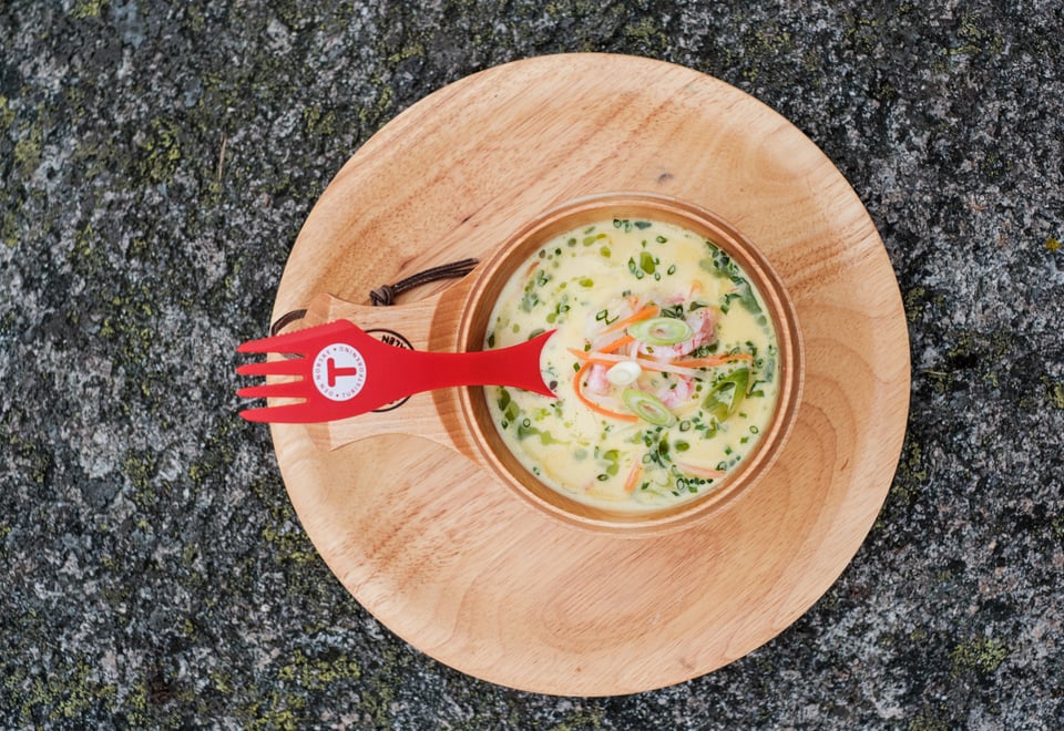 En tretallerken med en trekopp som inneholder fiskesuppe. Oppi suppa er det en rød skei med en T-logo på.