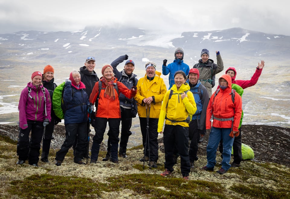 En turgruppe smiler og hoier på fjelltopp med skoddekledde fjell i bakgrunnen på høyfjellet.