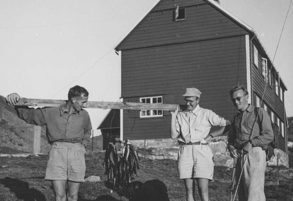 Gammelt bilde i svart-hvitt av tre menn som holder opp fisk foran hytte.