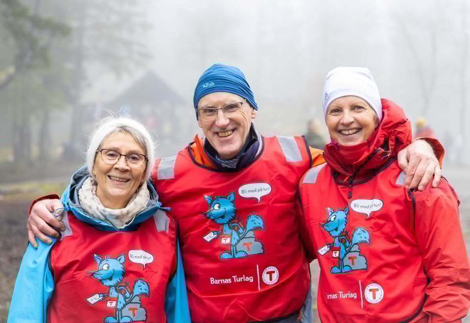 Tre frivillige med røde vester fra Turistforeningen smiler til kamera, en mann i midten som holder rundt to damer.