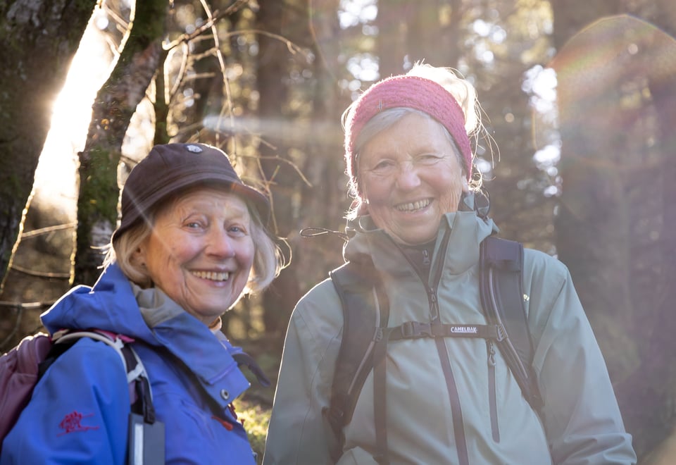 To voksne kvinner smiler til kamera når de er på tur i en skog.