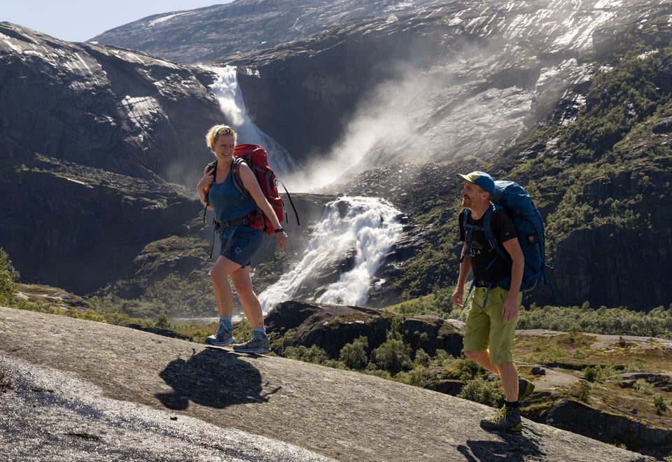 Kvinne på tur med mann. Hun snur seg mot ham og smiler med stor foss bak.