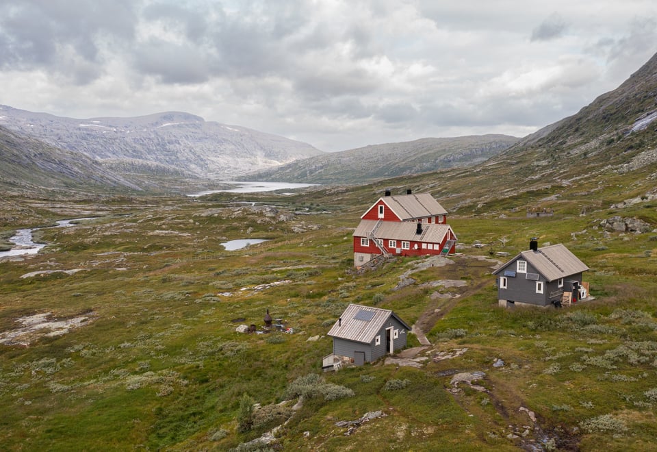 Rødmalt hytte i høyfjellet med to mindre blågrå hytter ved siden av.