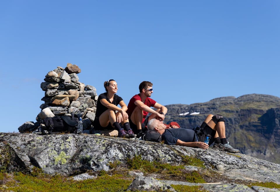 En kvinne og to menn sitter og slapper av ved en varde i solen på fjellet.