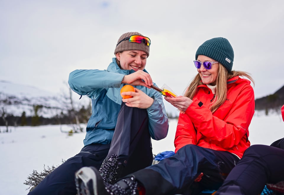 To kvinner sitter ute i vinterlandskap. De smiler og en holder en appelsin menst hun får sjokolade av den andre kvinnen. 