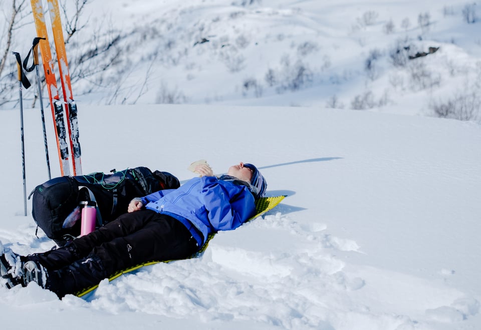 En person som ligger i snøen og soler seg. Det står et par med ski ved siden av