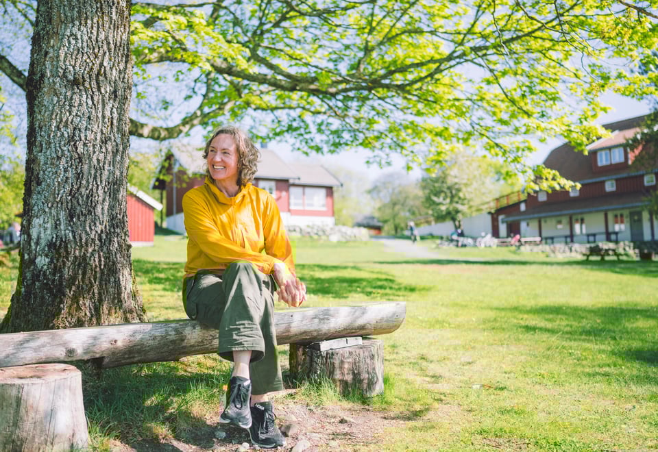 Kvinne i gult sittende på benk under et tre, grønt grass