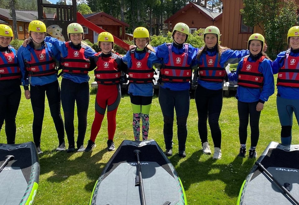 Ungdommer står på rekke foran paddleboards, de har på seg hjelm og redningsvest. 