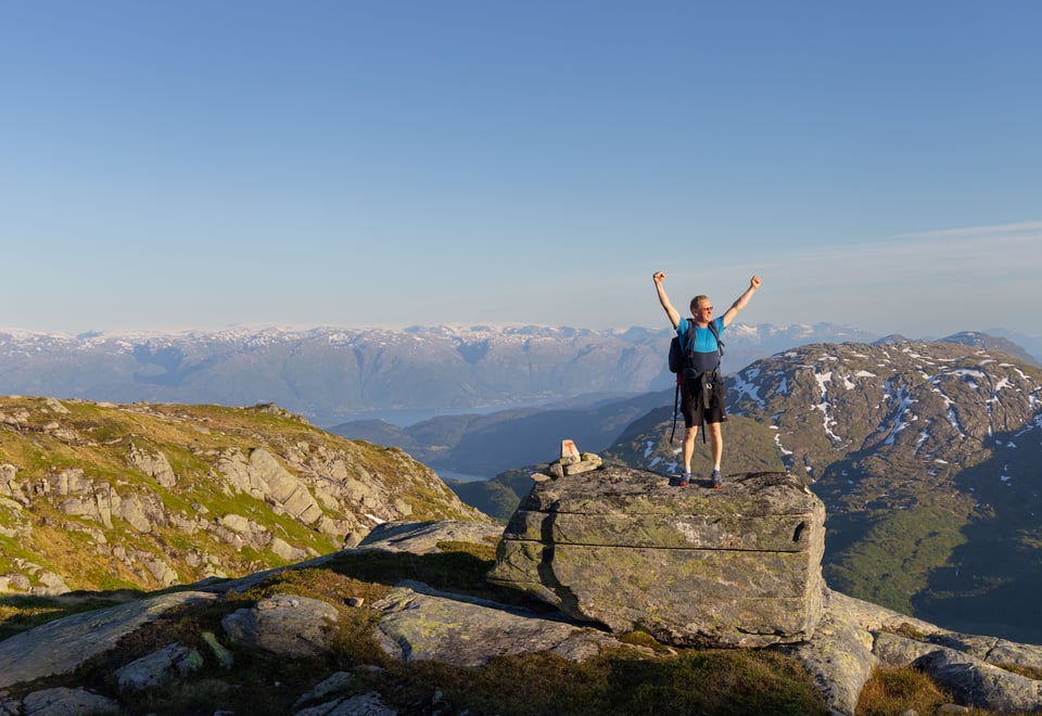 Mann 40+ står på stein med hendene i været, smiler, med Hardangerfjorden og fjell bak seg.