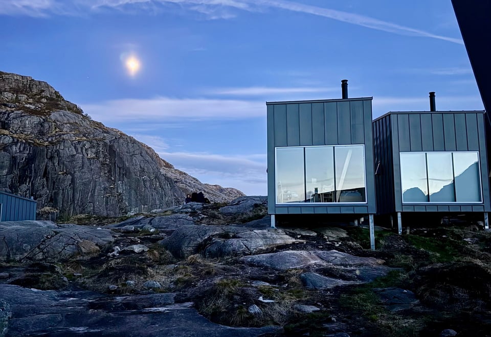 En grå hytte med store vinduer som ligger på fjellet. Måneskinn og kveldslys.