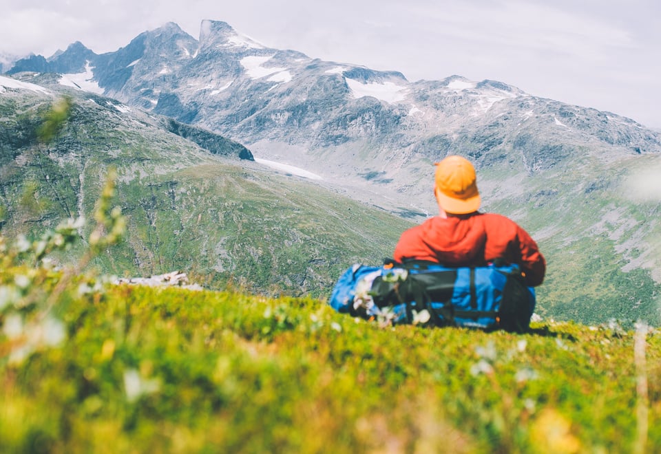 En mann i rød jakke og gul caps som sitter i lyngen og ser på fjell