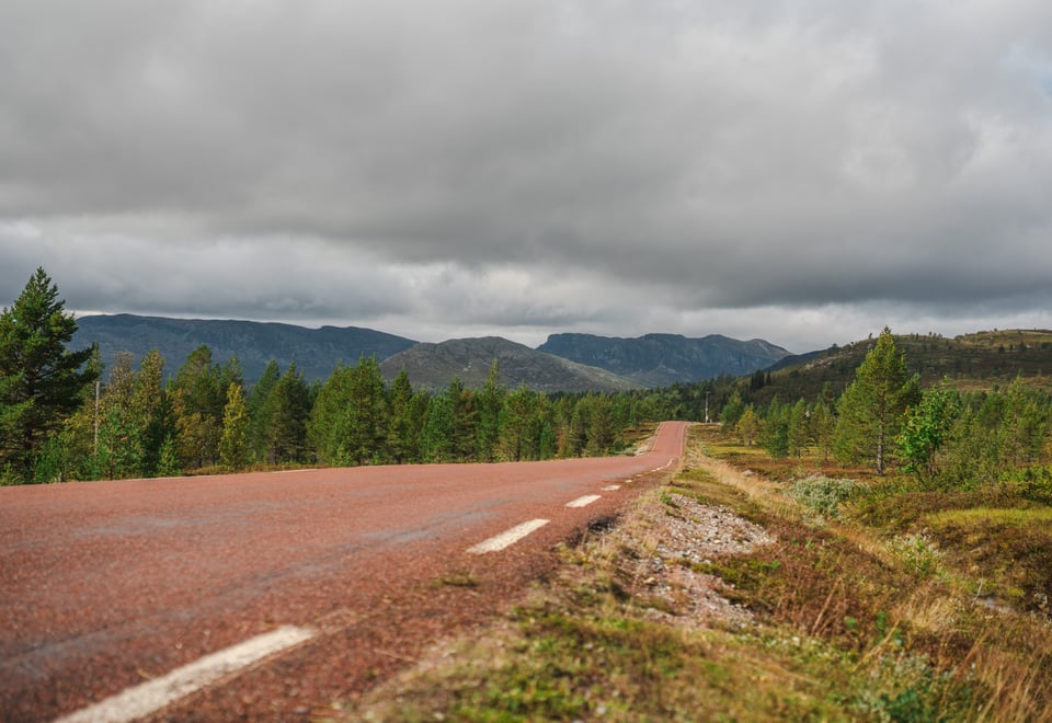 Rød asfaltert vei gjennom skogsområde, med fjell og grå skyer i bakgrunnen.