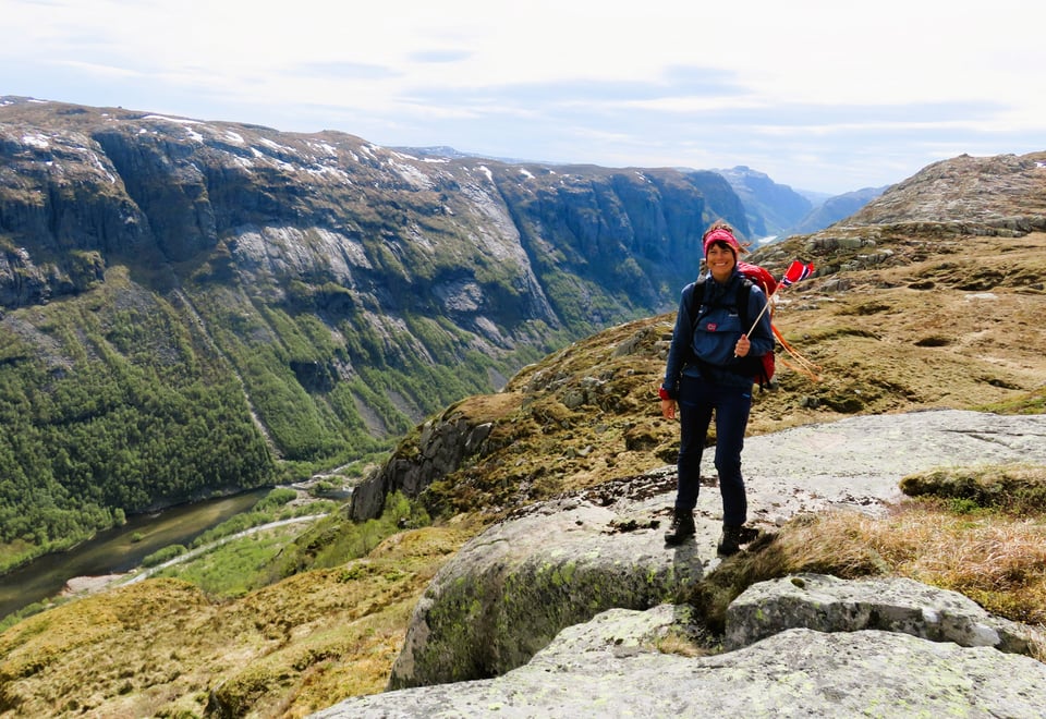 Kvinne med norsk flagg i hånden smiler til kamera, dalføre i bakgrunnen. 17. mai.
