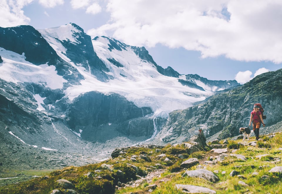 Dame med sekk og hund i bånd på sti med snødekte fjell i bakgrunnnen.