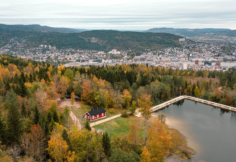 liten rød hytte som ligger i skogen ved et vann. i bakgrunnen ser man utsikt til en større by.