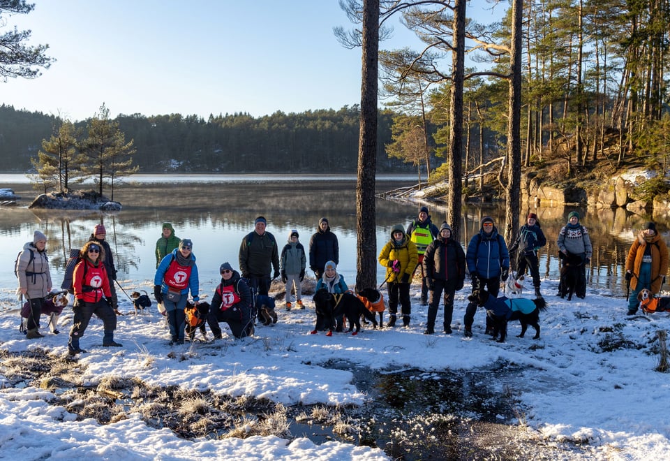 En gruppe menn og kvinner med hund står ved siden av hverandre foran et vann og snø i skogen.