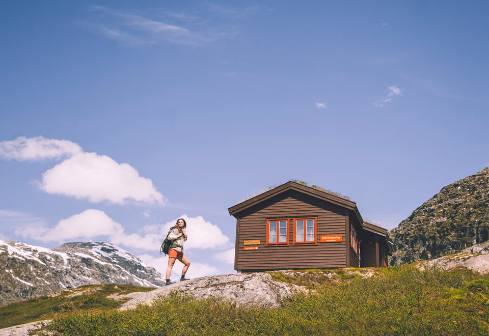 En person står foran en brun fjellhytte en sommerdag 