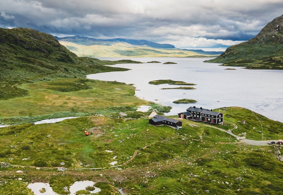 Iungsdalshytta sett ovenfra med drone en sommerdag, et fjellvann og en fjellkjede strekker seg utover horisonten