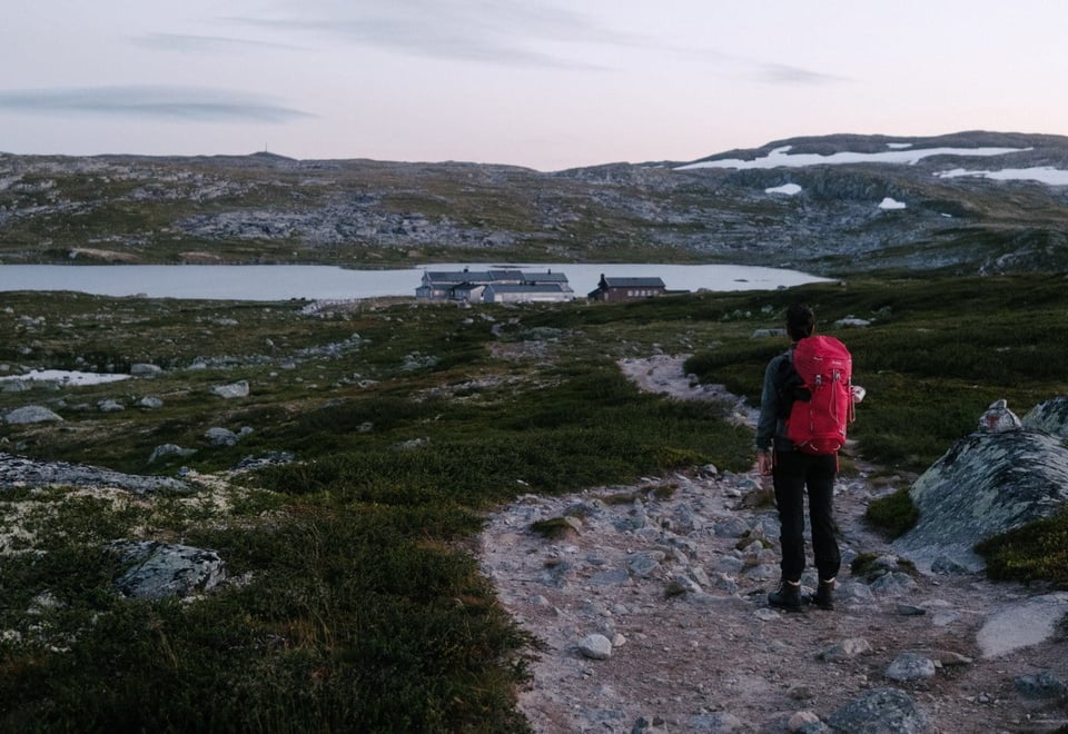 En person med rød sekk står foran en fjellhytte med et fjellvann i bakgrunnen