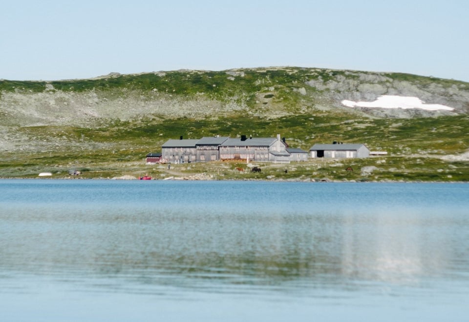 Et speilblankt fjellvann foran en lysegrå hytte i et fjellandskap 