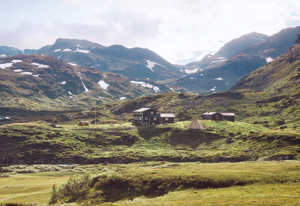En fjellhytta ligger i et vakkert sommerlandskap med grønne detaljer i forgrunnen, en elv og en fjellkjede bak