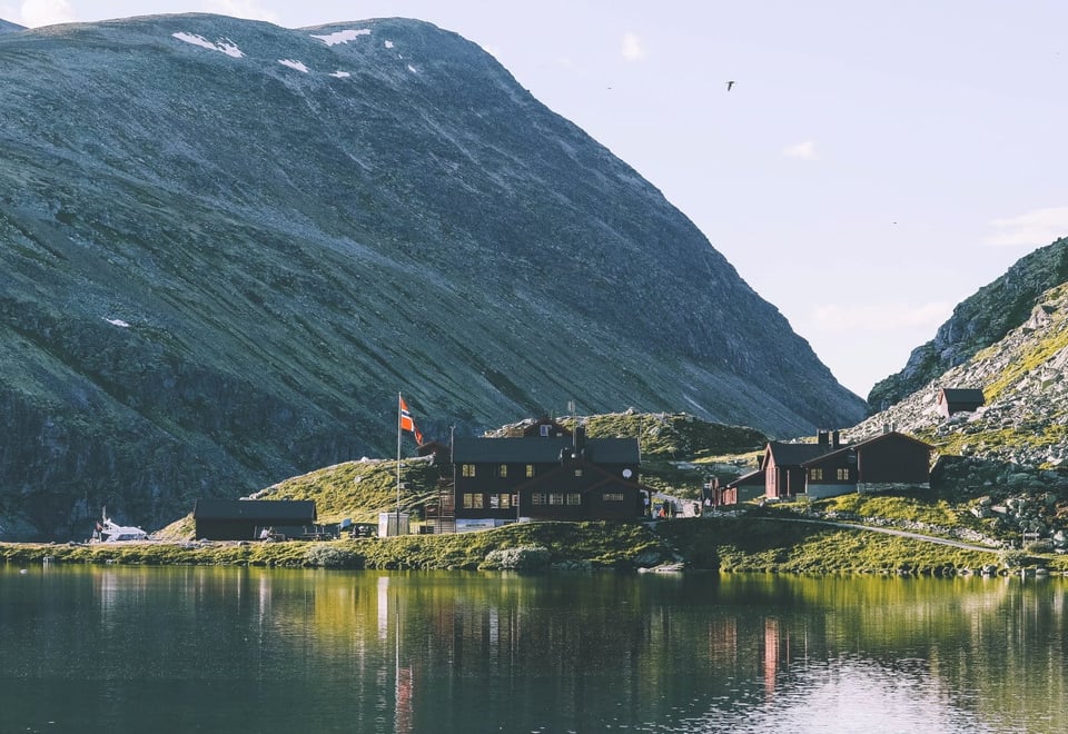 En fjellhytte med norskt flagg speiler seg i et fjellvann en sommerdag