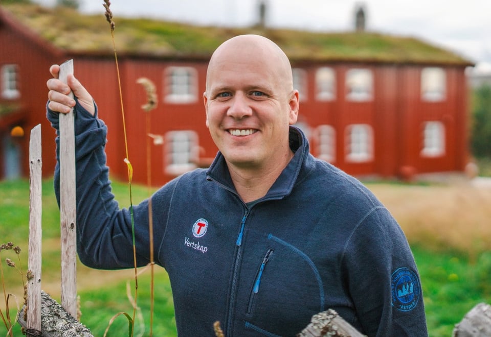 En person smiler til kamera foran en rød hytte