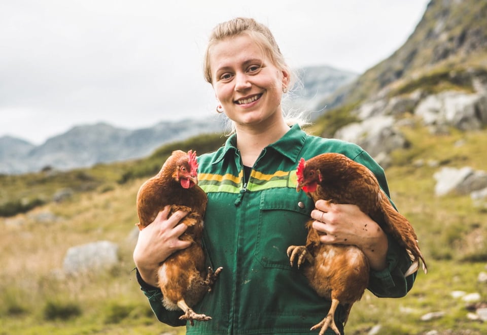 En dame i en grønn felleskjøpet dress - står smilende i et fjellandksap med to høner, en under hver arm 