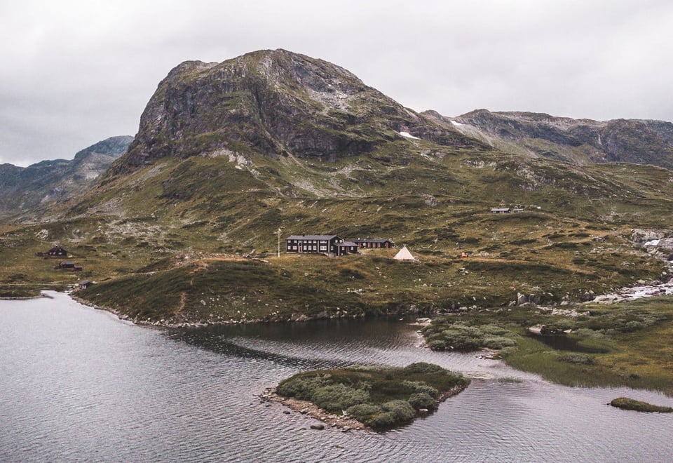 Et fjellandskap fotografert fra drone på sommeren med en liten hytte og et flott fjellvann i fogrunnen