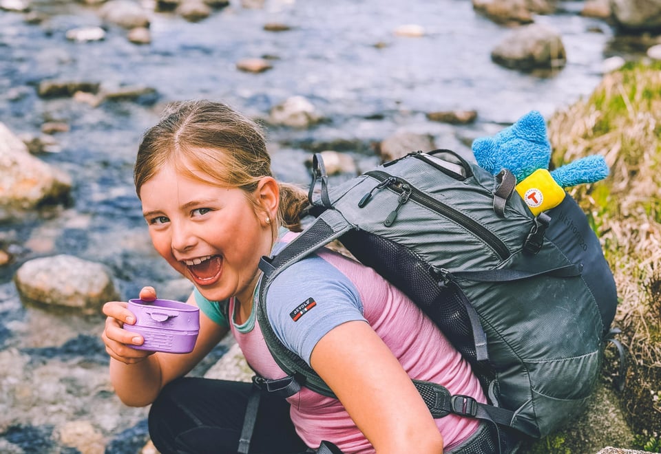 En liten jente sitter på huk og drikker fra en lilla kopp ved en elv. Hun smiler til kamera.