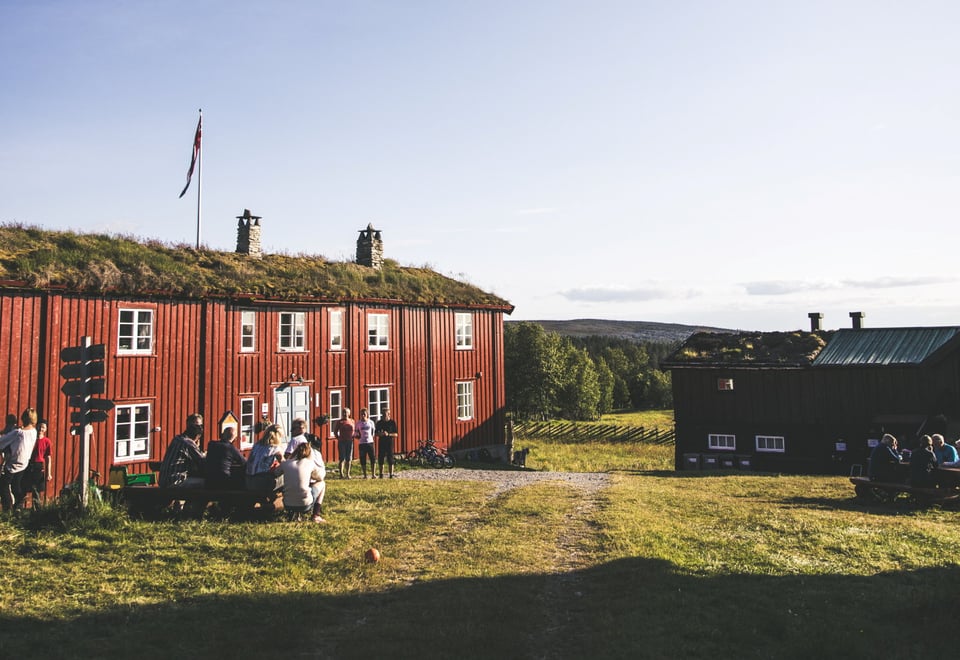 En gruppe mennesker sitter utenfor en rød hytte en sommersdag