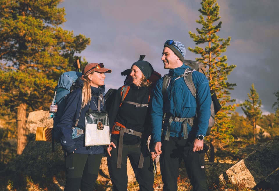 Tre turmennesker står i skogen og smiler til hverandre