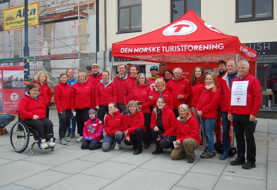 En gruppe mennesker med røde klær står foran en paviljong der det stor "Den norske turistforening"