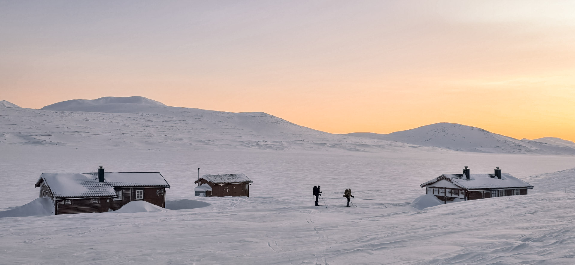 Hyttene i sn&oslash;landskap med en solnedgang bak dem. To mennesker p&aring; ski i bildet.