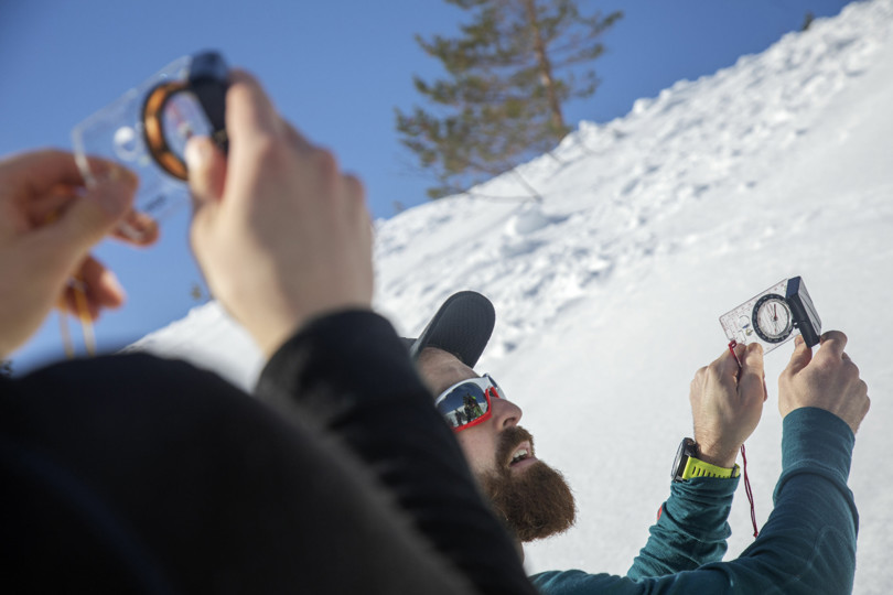 Mann med solbriller og caps holder opp et kompass for å måle helningsgraden på en snøkledd fjellside.
